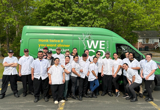 A group of smiling chefs and catering staff standing in front of their green catering van.