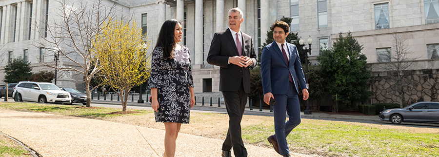 Two suited men and a woman in a dress walking down a path in a park.