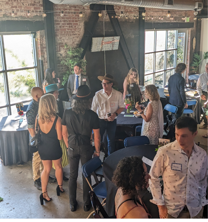 Guests mingling around cocktail tables at a networking event in a rustic industrial venue with exposed brick and large windows.
