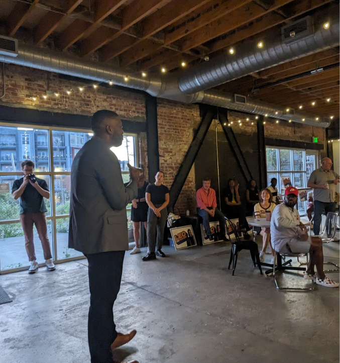 Speaker addressing a seated audience inside a brick-walled event space with large windows and string lights overhead.