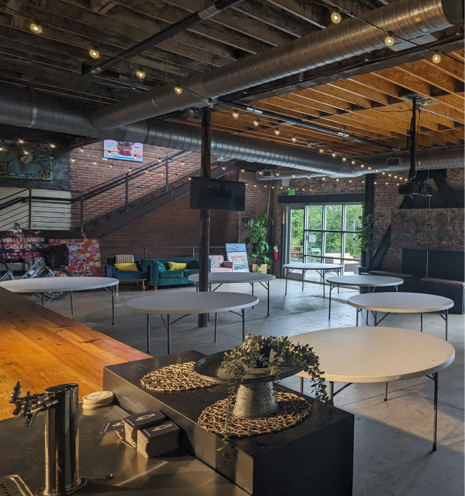 Crowd gathered at a networking event inside a modern loft-style venue with vendor tables, glass offices, and string lights.