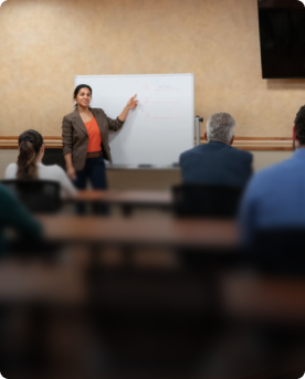 Instructor presenting at a whiteboard to a seated group in a large training room.