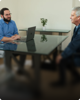Two professionals meeting at a glass desk in a private office, with a laptop open between them.