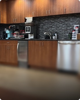 Modern office kitchen with wood cabinetry, coffee maker, and stocked beverage station.