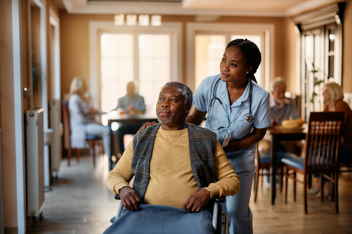 A man who uses a wheelchair looks off into the distance with a nurse, who places her hand on his shoulder and follows his gaze. In the background are other elderly residents at the facility.