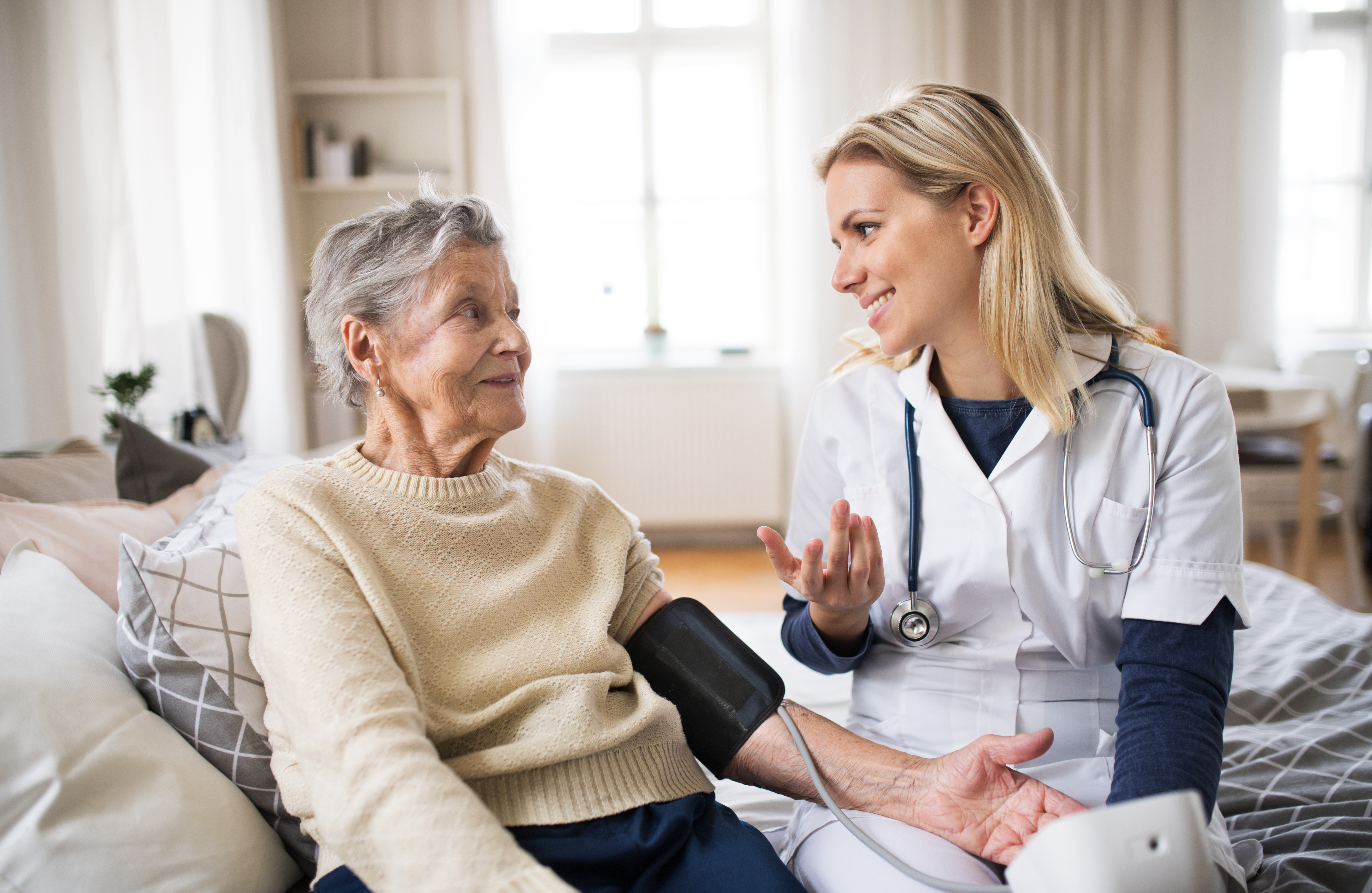 An elderly woman gets her blood pressure taken by a home nurse, who smiles at and enthusiastically speaks to her.