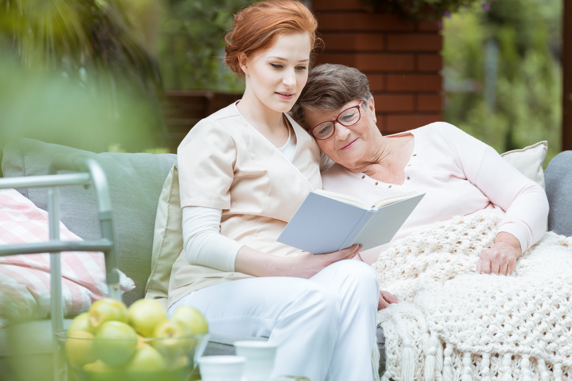 A nurse reads aloud to her patient, who leans her head on the nurse's shoulder and looks at the books pages. They are enjoying the outdoors.