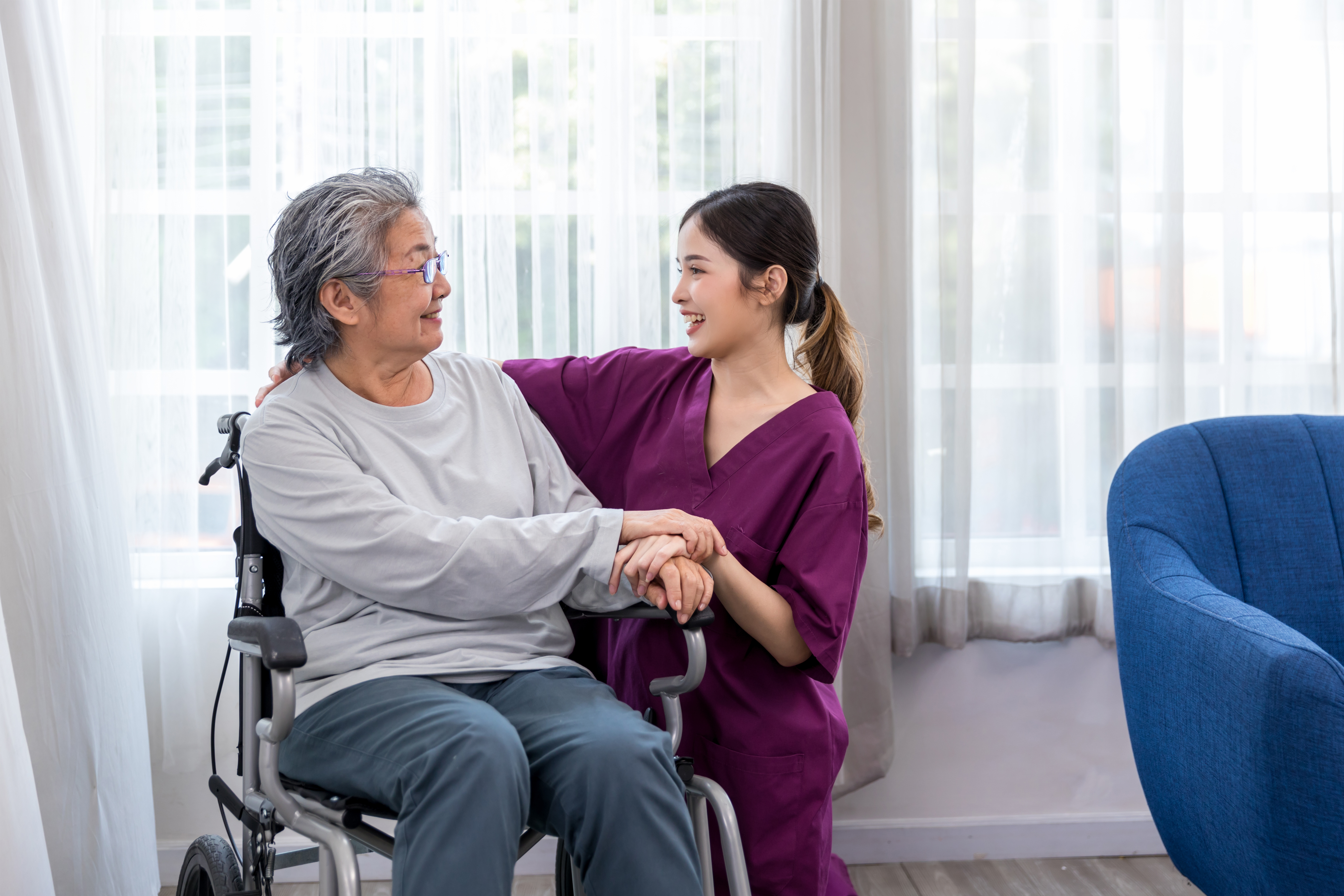 A wheelchair user is holding hands with and smiling at her nurse, who smiles back.