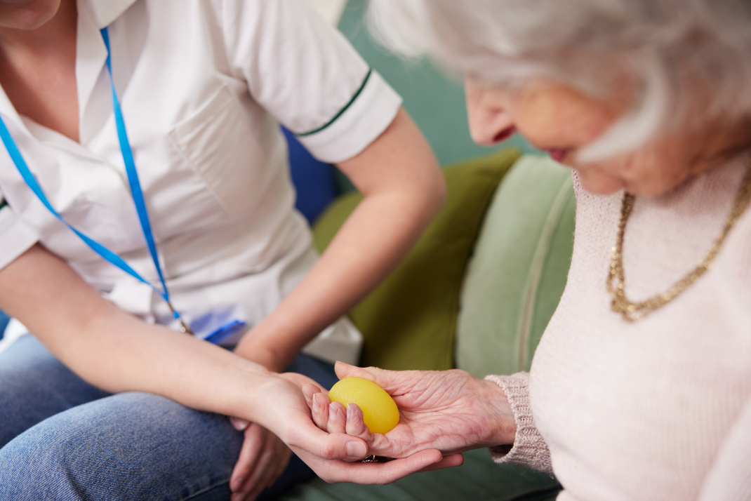 An elderly woman grasps a small plastic egg, being guided by the hand of an occupational therapist at her side.
