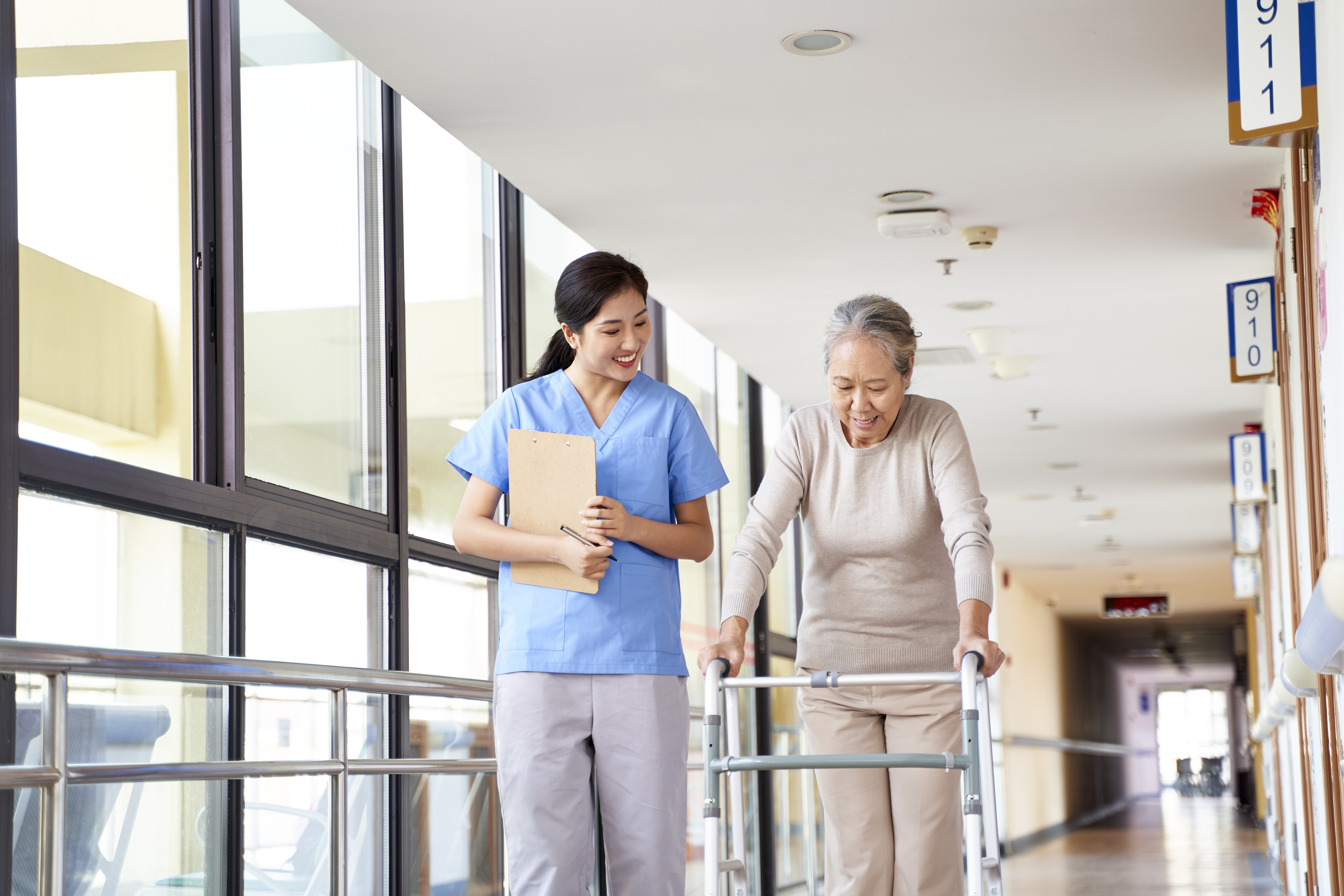 A nurse and her patient are in the hallway of a facility. The nurse looks at her patient, who uses a walker. They are both smiling. 