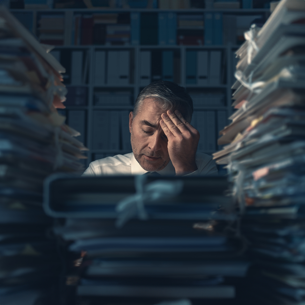 Stressed businessman with piles of paperwork in a cluttered office.