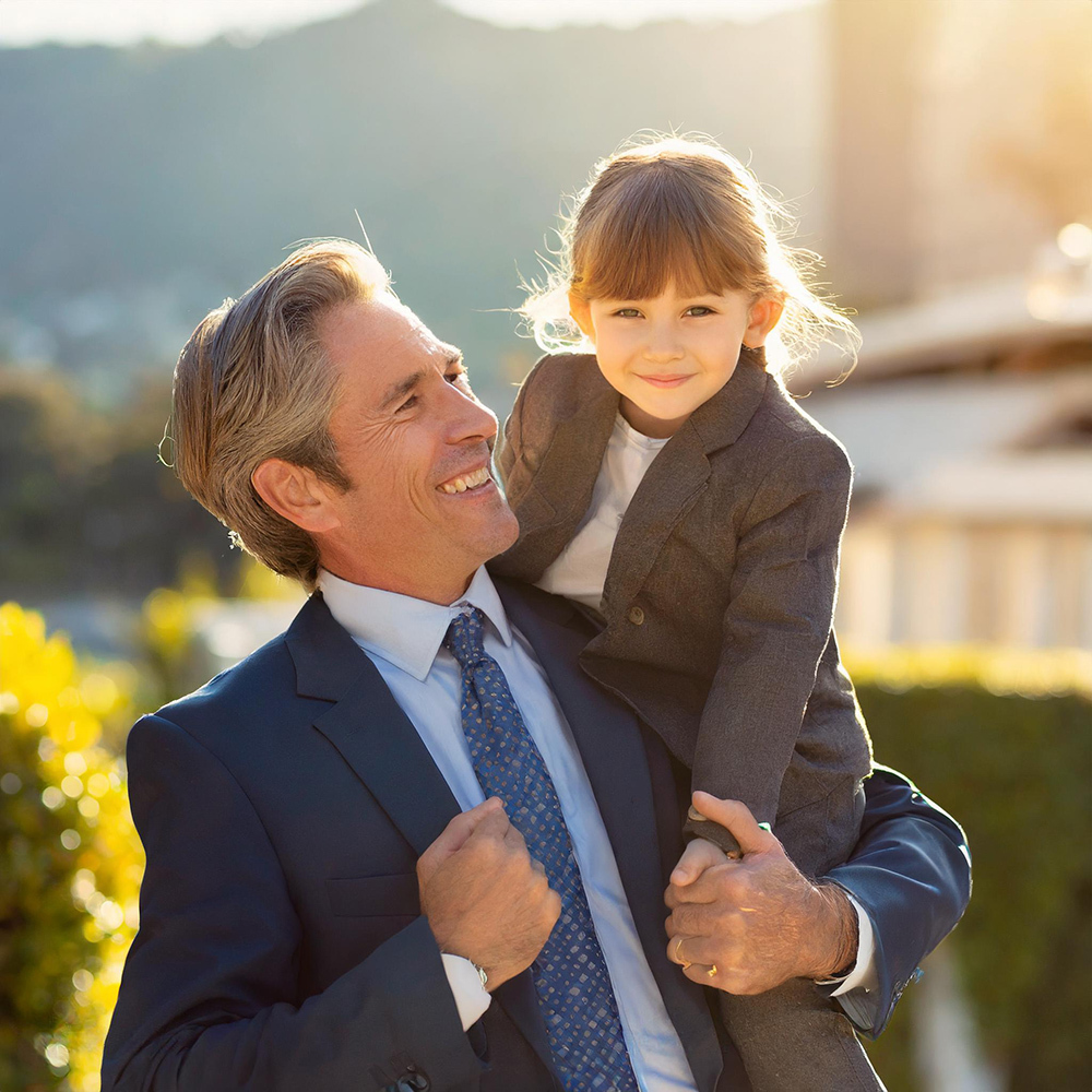 Smiling man in a business suit carrying a happy young girl on his shoulders outdoors