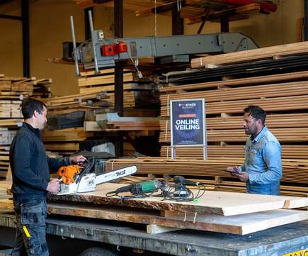Twee mannen in een houtwerkplaats met een kettingzaag en gereedschap op een werkbank, met een bord waarop 'Online Veiling' staat, representatief voor het veilen van bouwmachines en materialen