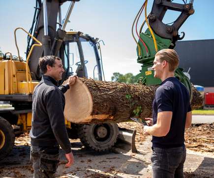 Twee mannen in gesprek naast een graafmachine met een boomstam in de grijper, representatief voor houtverwerking en het veilen van machines in de bouwsector.