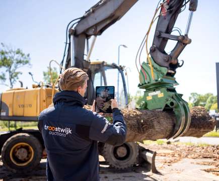 Medewerker van Troostwijk Auctions maakt een foto van een graafmachine met grijper die een boomstam vasthoudt, representatief voor het veilen van bouw- en constrcutiemachines