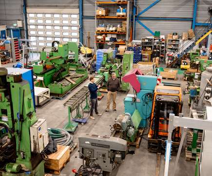 Overzicht van een werkplaats met diverse metaalbewerkingsmachines en apparatuur, met twee mannen in gesprek over verkoop, representatief voor de metaalbewerking en industriële veilingen.